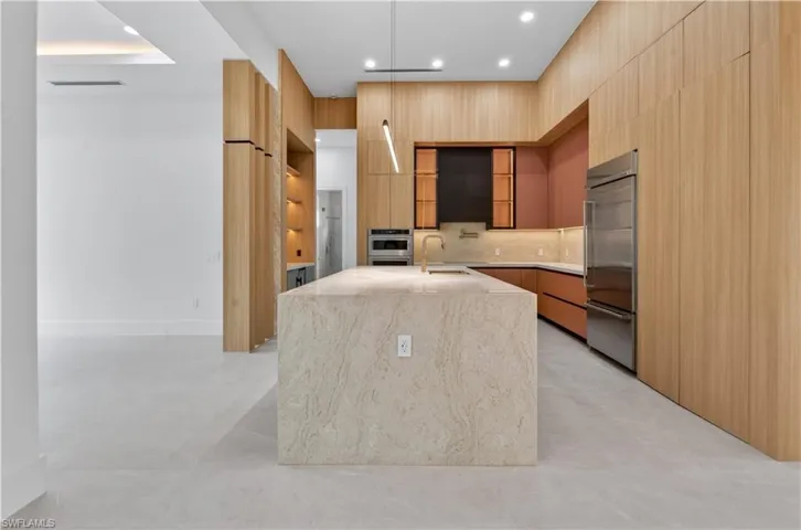Modern kitchen featuring a large stone-finish island with integrated sink and gold-tone faucet, light wood-finish cabinetry, stainless steel refrigerator, and recessed lighting