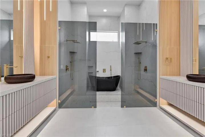 Contemporary bathroom featuring twin walk-in showers with clear glass enclosures and brass fixtures, a freestanding black soaking tub, and dual floating vanities with reeded cabinetry and vessel sinks