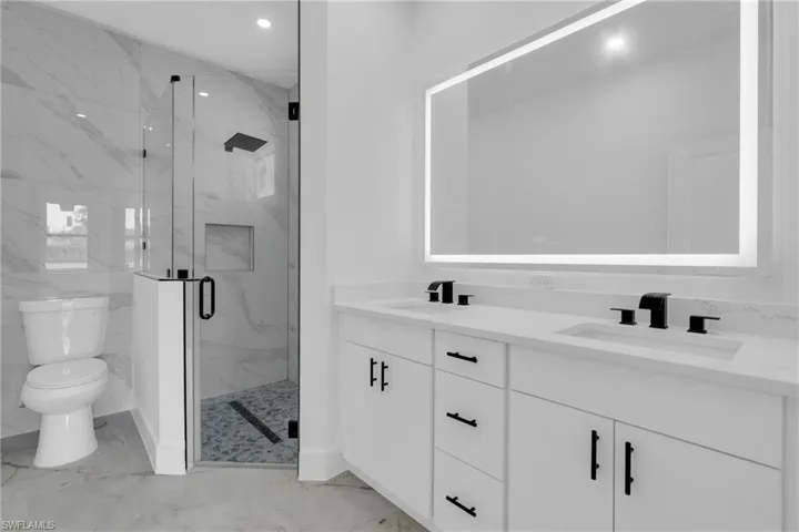 Dual vanity with matte black fixtures and white countertops, illuminated mirror, glass-enclosed shower with a black shower head, and large format wall tile
