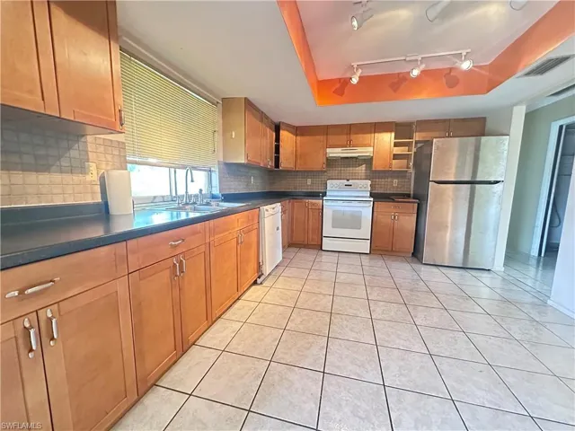 Kitchen with white appliances, a sink, under cabinet range hood, dark countertops, and tasteful backsplash
