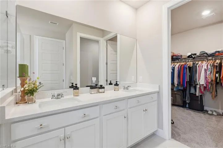 Dual vanity with undermount sinks, white cabinetry, expansive mirror, and a walk-in closet with built-in shelving and carpet flooring