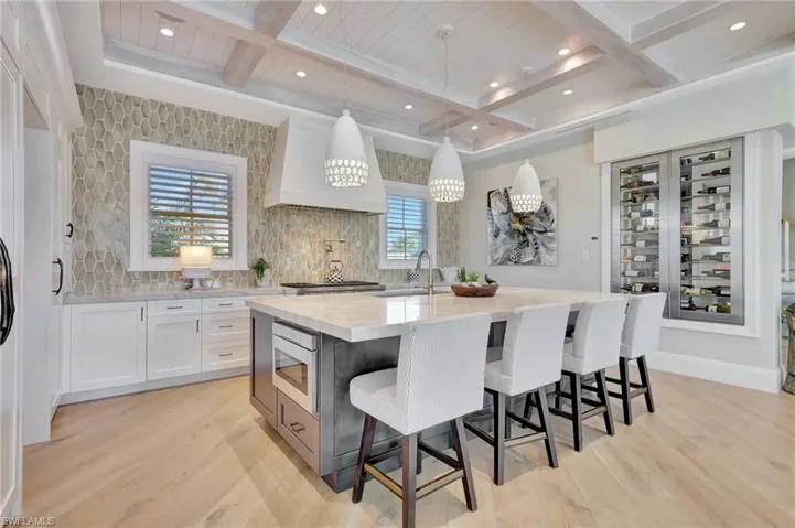 Kitchen, a center island with sink, unique pendant lighting, light wood flooring, white cabinetry, and enclosed wine wall