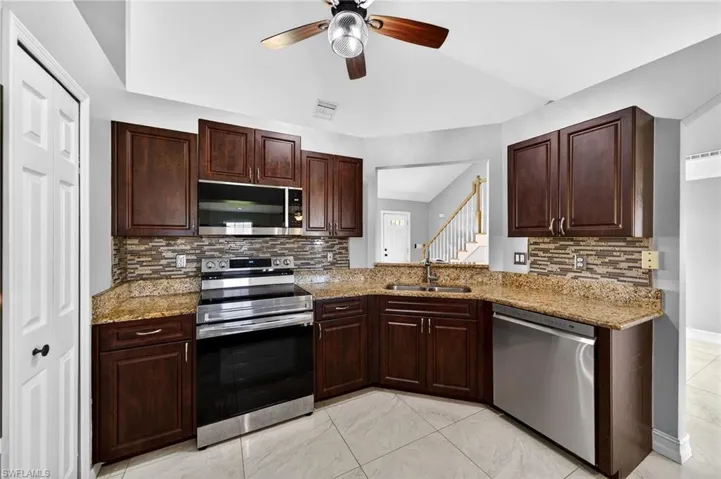 Kitchen with stainless steel appliances, dark wood finish cabinets, light stone countertops, ceiling fan, and vaulted ceiling