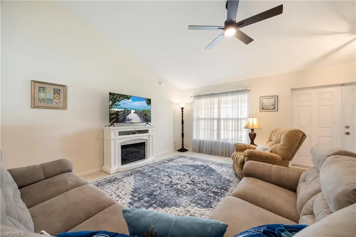 Vaulted ceiling living area featuring a decorative electric fireplace, an overhead ceiling fan with lighting, large front windows, and tile flooring