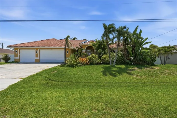 Yellow stucco exterior featuring a terra cotta tile roof, two white garage doors, a concrete driveway, and established tropical landscaping