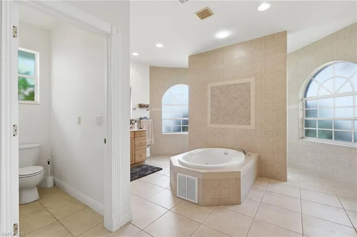 Spacious bathroom featuring a built-in jetted tub, arched windows with decorative grilles, and neutral ceramic tile flooring