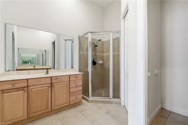Spacious bathroom featuring a wood-finish vanity with a light-toned countertop, a large wall-mounted mirror, a tiled corner shower with a glass enclosure, and light-toned tile flooring