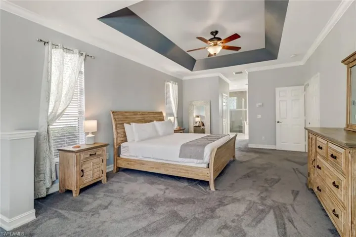 Carpeted bedroom featuring crown molding, a raised ceiling, ensuite bath, ceiling fan, and multiple windows