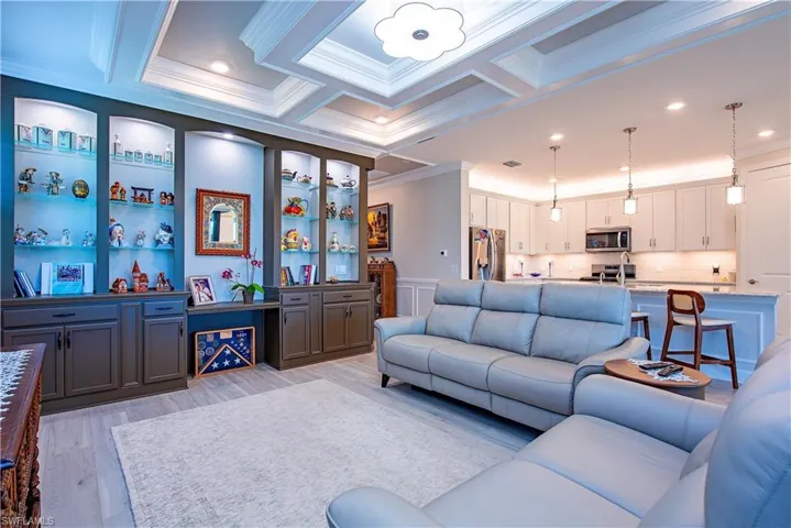 Open-concept living area featuring custom built-in cabinetry with illuminated glass shelving, coffered ceiling, and wood-finish flooring