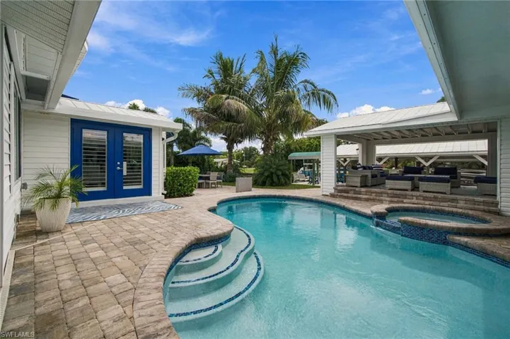 Outdoor swimming pool with built-in spa, expansive paver patio, covered outdoor living area, and bright blue French doors