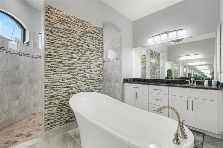 The bathroom features a freestanding soaking tub, a tiled shower with an arched window, and a dual vanity with a dark countertop and white cabinetry