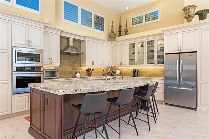 Kitchen with built in appliances, glass insert cabinets, light stone countertops, a breakfast bar, and wall chimney range hood