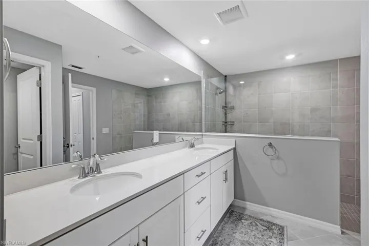 Dual-sink vanity with white cabinetry, light-toned solid surface countertop, full-width vanity mirror, and walk-in shower featuring tiled wall surround