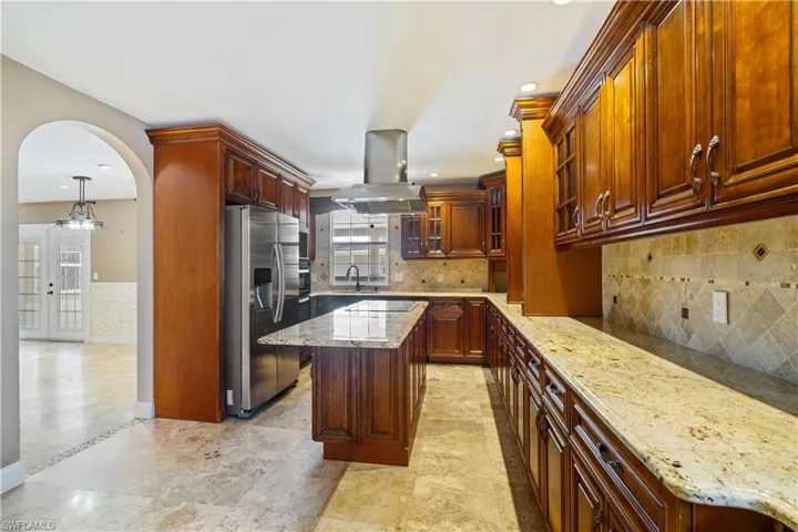 Kitchen with light granite countertops, glass insert cabinets, stainless steel refrigerator with ice dispenser, island exhaust hood, and arched walkways