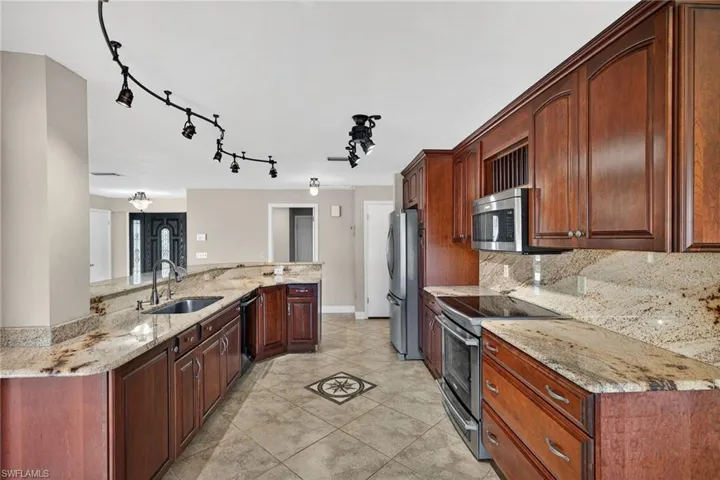 Custom wood finished cabinetry featuring Electrolux appliances with stainless steel finishes, light stone countertops, rail lighting, light tile patterned flooring, and decorative backsplash