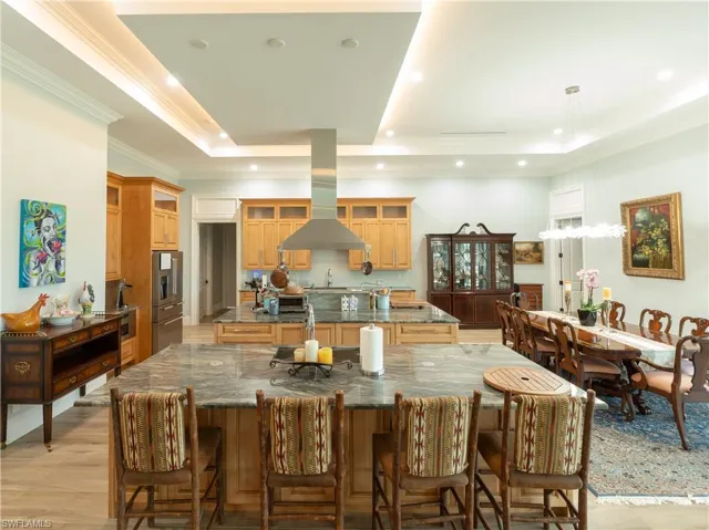 Kitchen with a large island with sink, island exhaust hood, a raised ceiling, glass insert cabinets, and recessed lighting