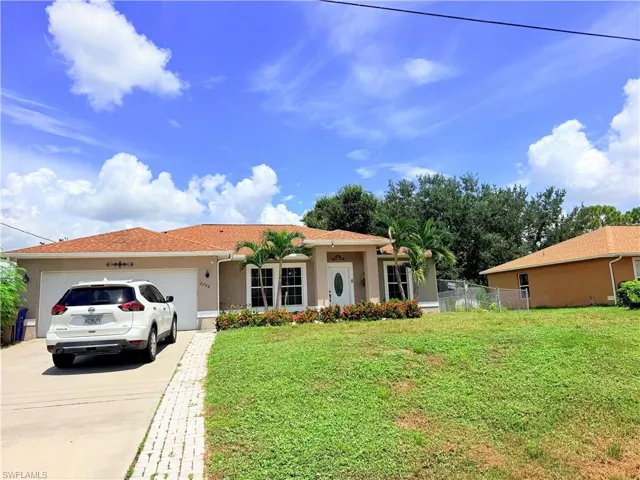 Concrete driveway with brick inlay, attached single-car garage, stucco exterior, front landscaping, and a chain-link fence