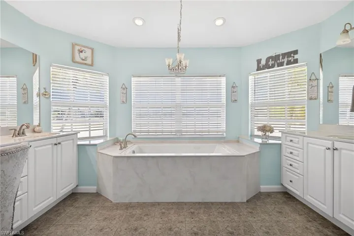 Recessed soaking tub with surrounding windows, dual vanity sinks with white cabinetry, and a central chandelier