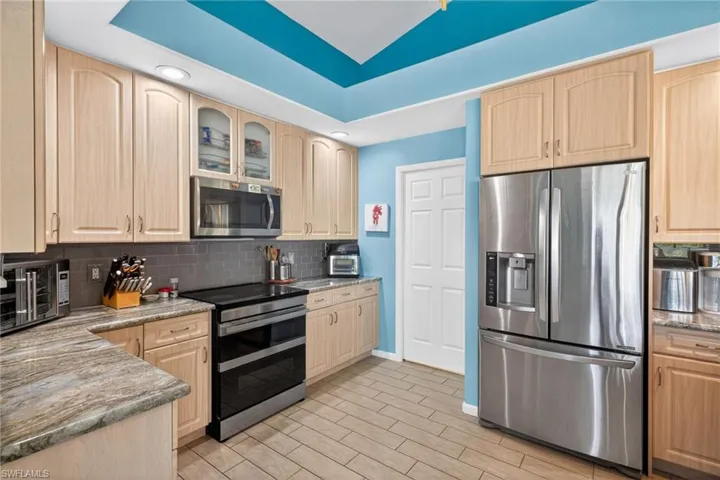 Kitchen featuring light wood-finish cabinetry, stainless steel appliances, a tile backsplash, stone-finish countertops, and wood-look tile flooring