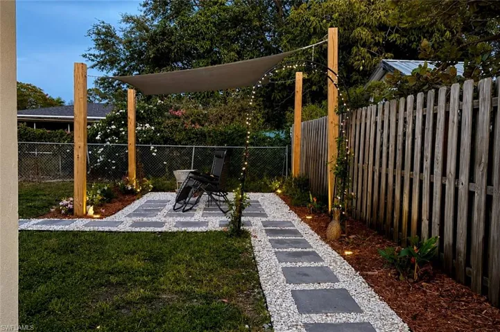 Backyard patio area featuring a paver stone and white gravel walkway, a wooden privacy fence, a shade sail supported by wooden posts, and integrated ground lighting