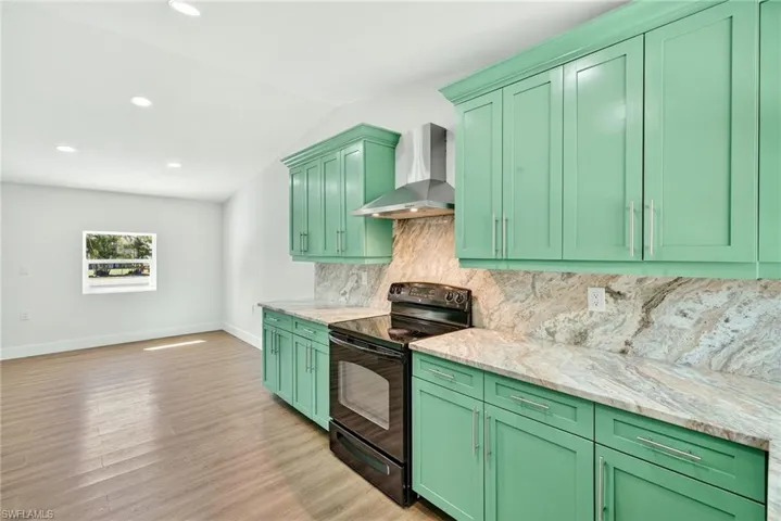 Kitchen with green cabinetry, black / electric stove, wall chimney range hood, light stone countertops, and tasteful backsplash
