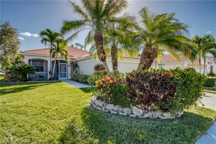 Exterior facade featuring a terracotta-style tile roof, stucco finish, stone-lined garden bed, mature palm trees, and a covered entryway