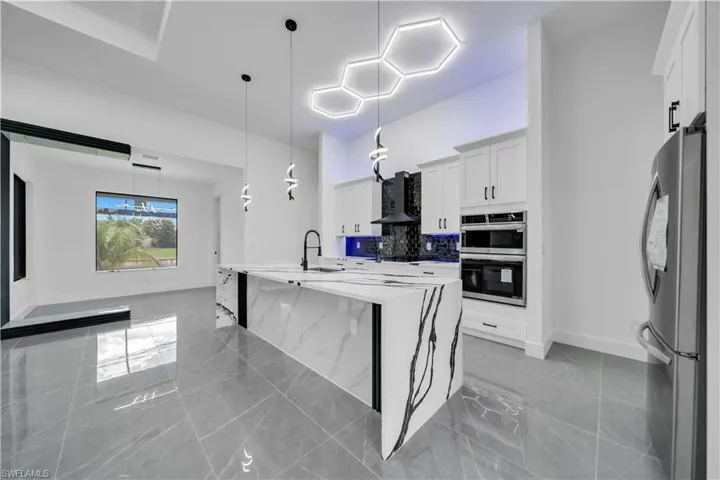 Kitchen featuring appliances with stainless steel finishes, wall chimney range hood, white cabinetry, light marble finish flooring, and pendant lighting