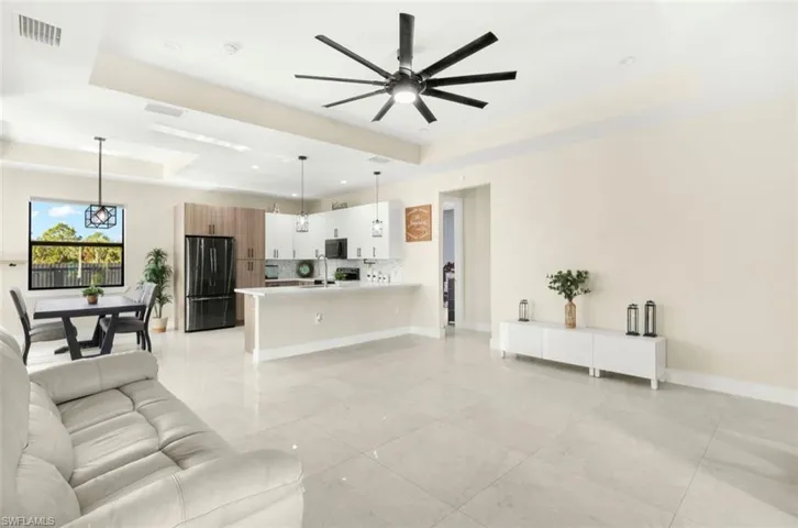 Open-concept living space with large format tile flooring, a contemporary kitchen featuring white cabinetry and black appliances, a dining area with pendant lighting, and a tray ceiling with a modern ceiling fan