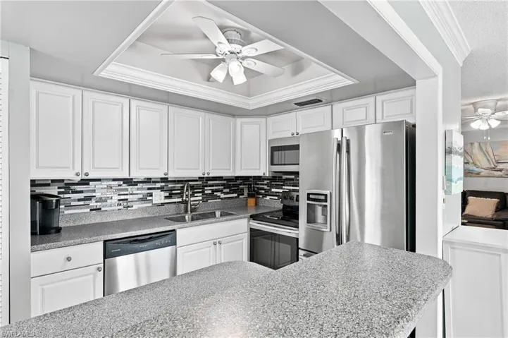 White cabinetry, stainless steel appliances, mosaic tile backsplash, recessed ceiling with crown molding, and a ceiling fan