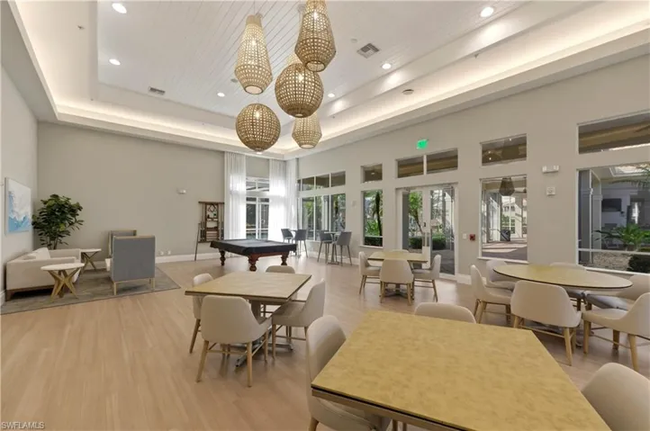 Spacious common area featuring light wood-finish flooring, a high ceiling with recessed lighting and decorative woven pendants, a full wall of windows and glass doors, and a billiards table