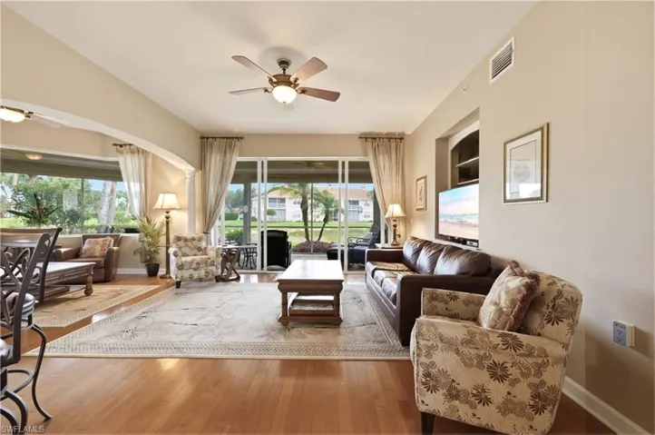 Living area with wood-finish flooring, featuring a ceiling fan with integrated lighting, a recessed wall niche with shelving, and sliding glass doors opening to an outdoor screened area