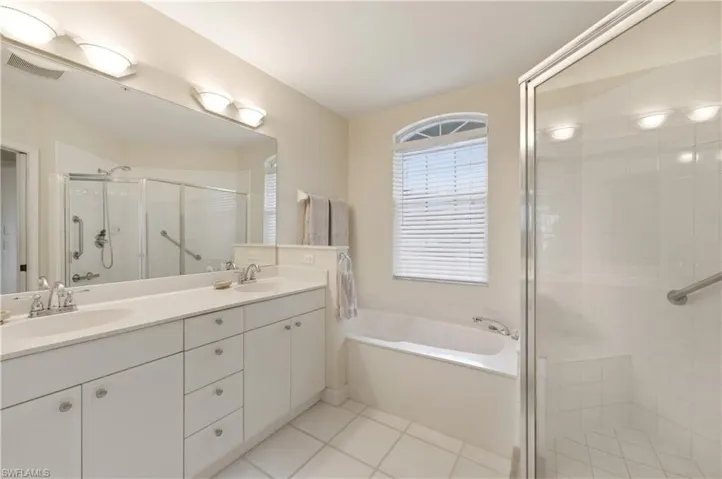 Spacious bathroom featuring a dual vanity with white cabinetry, a large frameless mirror, a built-in soaking tub, a separate glass-enclosed shower, and a window with arched transom