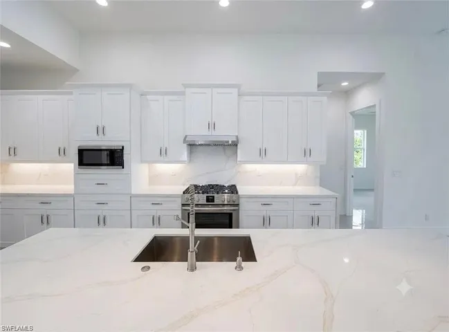 Kitchen featuring white cabinetry, a gas range with a stainless steel hood, built-in microwave, a stainless steel sink, and light-toned countertops with veining