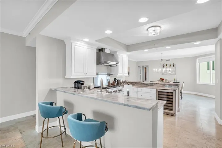 Kitchen with wine cooler, white cabinets, kitchen peninsula, wall chimney range hood, and light stone countertops