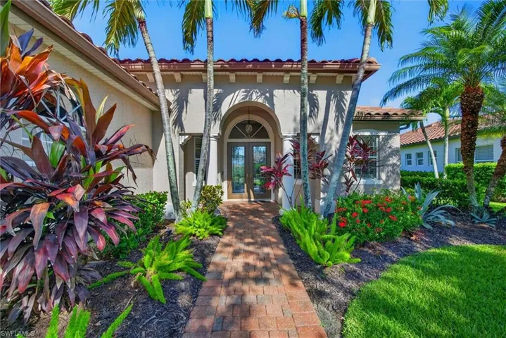 Inviting entryway featuring a brick paver pathway, lush tropical landscaping with varied foliage, a prominent arched double-door entry, exterior stucco finish, and a tile roof