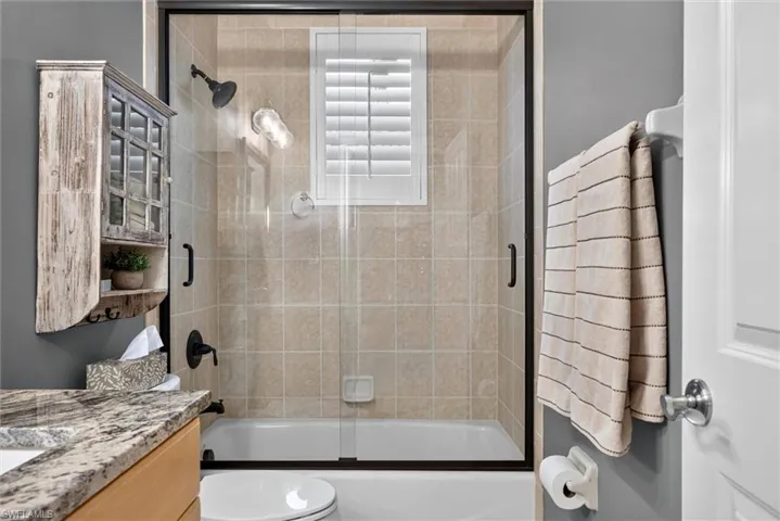 Bathroom featuring a tiled shower/tub combination with a sliding glass door, a built-in window with horizontal blinds, a vanity with a stone countertop, and a light wood-finish cabinet