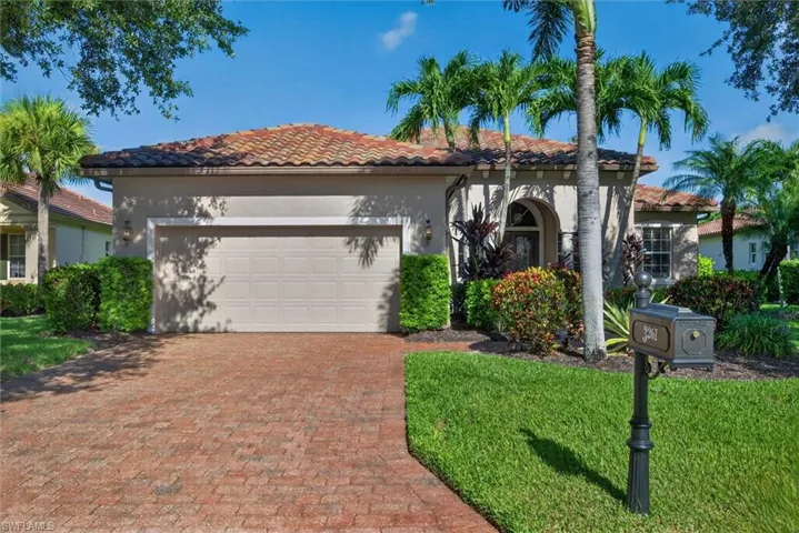 Stucco exterior featuring a barrel tile roof, brick paver driveway, an arched entry, mature landscaping, and an attached garage