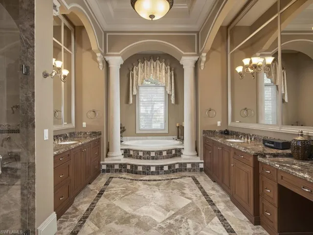 Full bathroom with inlaid floor details, two vanities, a garden tub, ornate columns, and ornamental molding