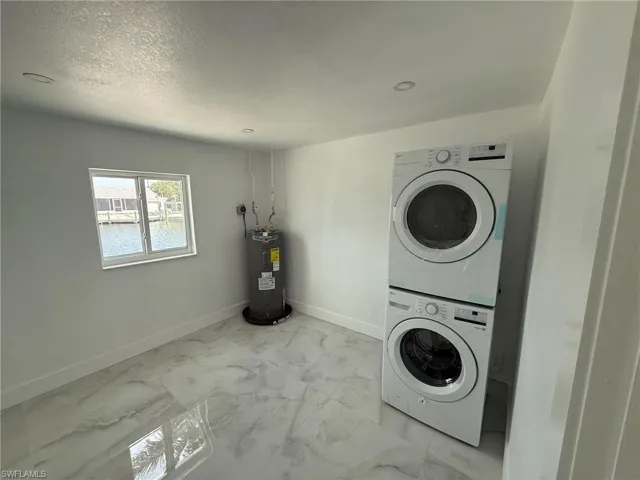 Laundry area featuring light marble finish flooring, stacked washer / dryer, a textured ceiling, and electric water heater