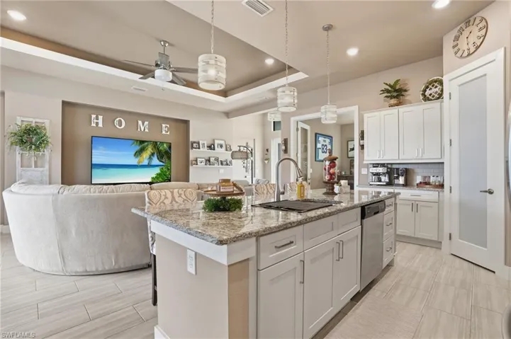 Kitchen with ceiling fan, open floor plan, light stone counters, an island with sink, and a raised ceiling