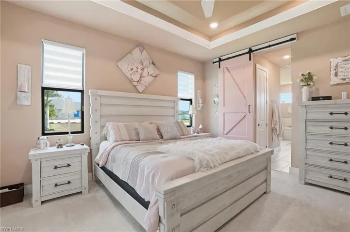 Bedroom with a barn door, multiple windows, light colored carpet, a tray ceiling, and a ceiling fan
