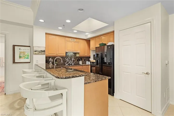 Kitchen featuring dark stone counters, kitchen peninsula, black appliances, backsplash, and light tile floors