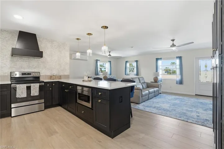 Kitchen featuring stainless steel appliances, a peninsula, wall chimney exhaust hood, open floor plan, and decorative light fixtures - Virtually Edited Image