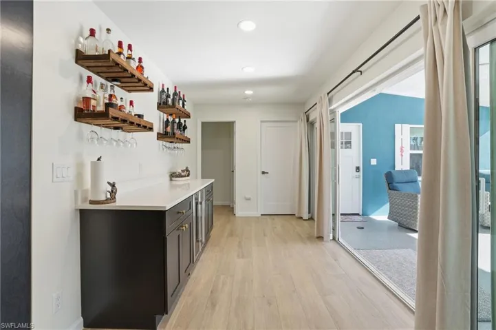 Indoor dry bar featuring light wood tile floors, open shelves, recessed lighting, dark cabinets, and light stone counters - Virtually Edited Image