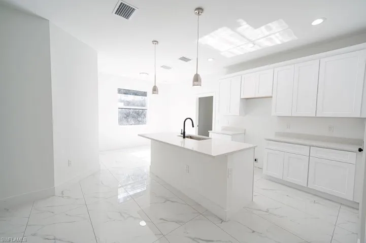 Kitchen featuring a kitchen island with sink, white cabinetry, hanging light fixtures, light stone counters, and light marble finish flooring. Photos are of a staged model or similar plan. Features and finishes may vary.