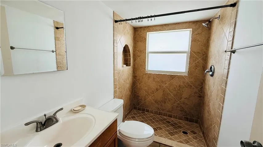 Bathroom featuring a fully tiled shower enclosure with a window, decorative arched shower niche, white toilet, and a vanity with a white countertop and dark-toned faucet