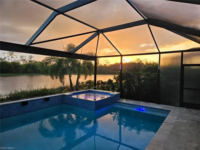 Pool at dusk featuring a water view, glass enclosure, and a pool with connected hot tub