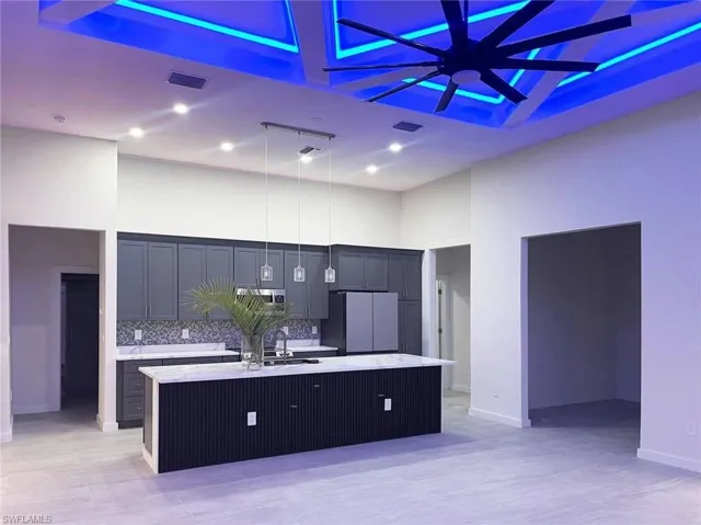 Open-concept kitchen featuring an oversized island with a white countertop and pendant lighting, dark gray cabinetry, stainless steel appliances, and a mosaic tile backsplash