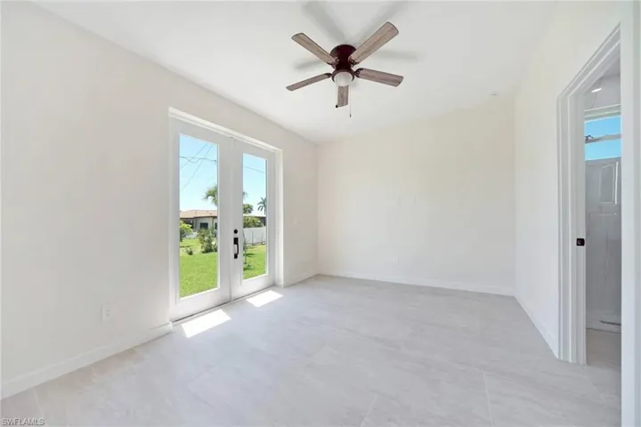 Spacious room featuring light-toned flooring, contemporary ceiling fan, white baseboards, and double glass doors opening to a lush green lawn