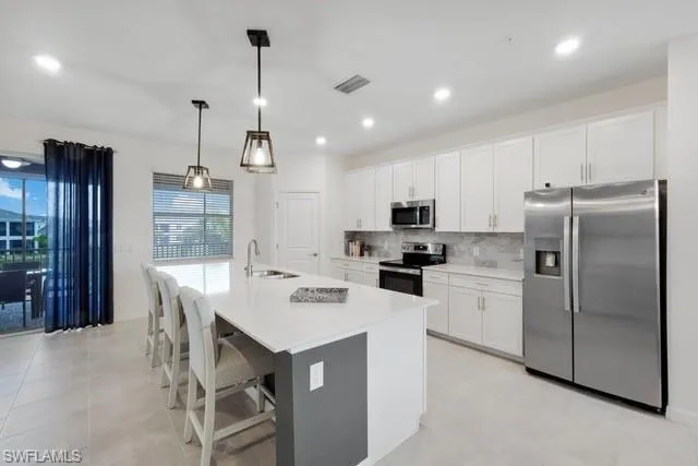 Kitchen featuring appliances with stainless steel finishes, a sink, backsplash, a kitchen island with sink, and white cabinets